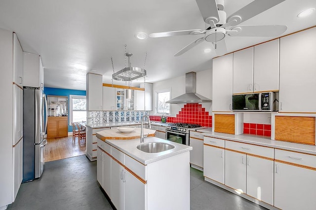 kitchen with a center island with sink, a sink, backsplash, stainless steel appliances, and wall chimney exhaust hood