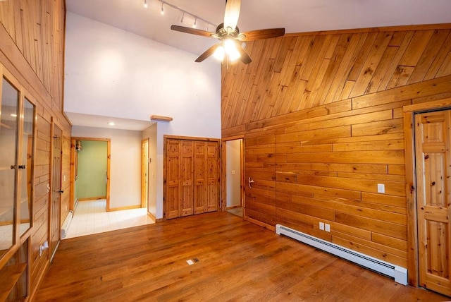 unfurnished room with wooden walls, wood finished floors, high vaulted ceiling, a baseboard radiator, and ceiling fan