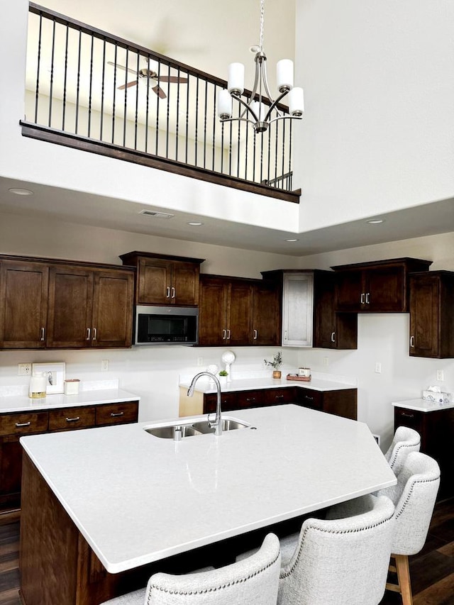 kitchen featuring a center island with sink, a sink, dark brown cabinetry, a towering ceiling, and stainless steel microwave