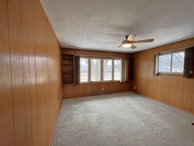 carpeted spare room featuring built in features, wood walls, baseboard heating, and ceiling fan