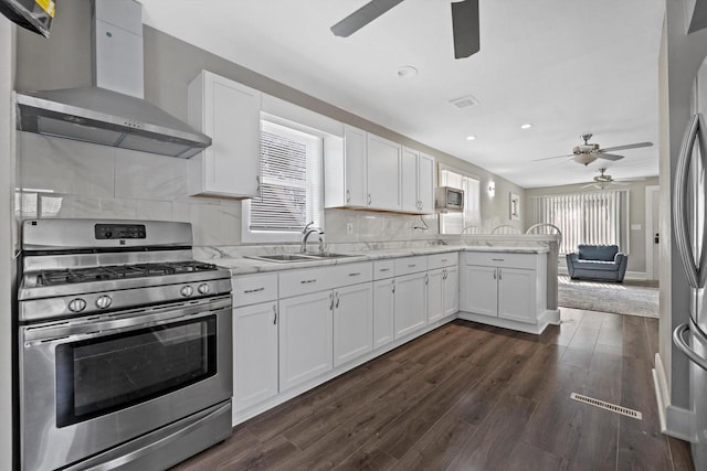 kitchen with wall chimney range hood, a peninsula, white cabinets, stainless steel appliances, and a sink