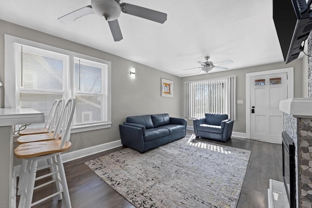 living area with dark wood-style floors, a stone fireplace, a ceiling fan, and baseboards