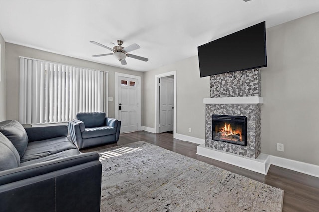 living area with a stone fireplace, a ceiling fan, dark wood-type flooring, and baseboards