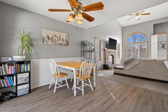 dining area with a brick fireplace, ceiling fan, a wainscoted wall, vaulted ceiling, and wood finished floors