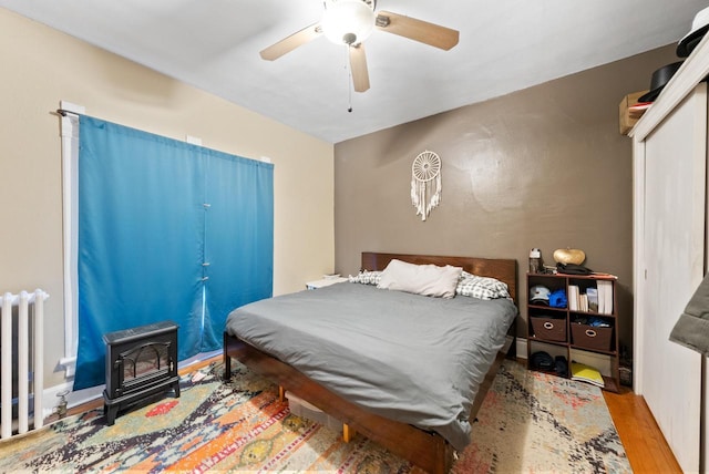 bedroom featuring a ceiling fan, a wood stove, radiator heating unit, and wood finished floors