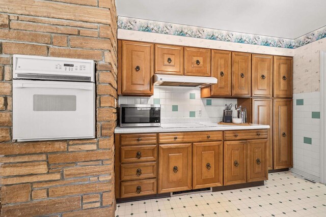 kitchen featuring brown cabinetry, wallpapered walls, under cabinet range hood, white oven, and stainless steel microwave