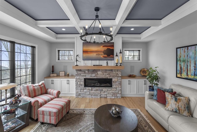 living area with coffered ceiling, beam ceiling, light wood-style flooring, a stone fireplace, and a chandelier