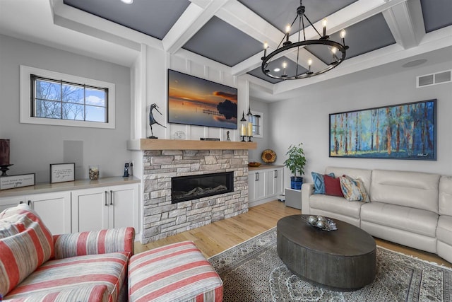 living room with visible vents, light wood-type flooring, a stone fireplace, a notable chandelier, and coffered ceiling