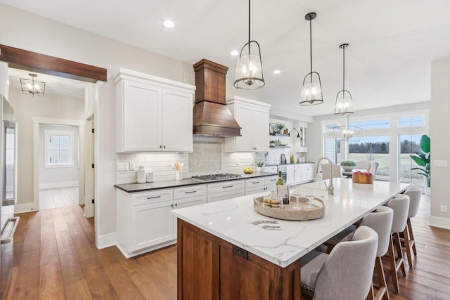 kitchen with a kitchen island with sink, custom range hood, white cabinets, decorative backsplash, and stainless steel gas cooktop