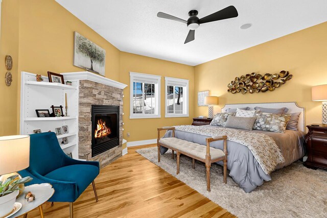 bedroom with a ceiling fan, a textured ceiling, wood finished floors, a stone fireplace, and baseboards