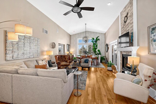 living room featuring light wood finished floors, visible vents, a stone fireplace, ceiling fan with notable chandelier, and high vaulted ceiling