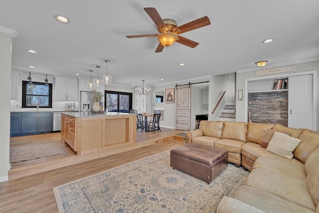 living area with a barn door, light wood-style floors, crown molding, and ceiling fan with notable chandelier