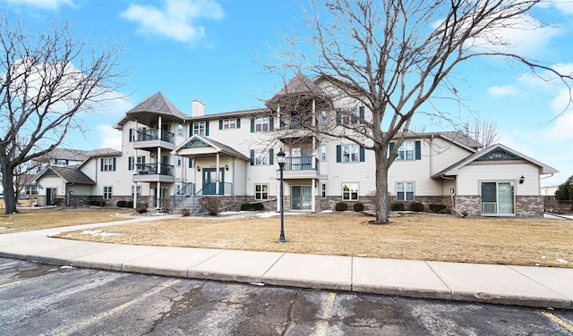 view of property with a front lawn, a chimney, a balcony, stone siding, and uncovered parking