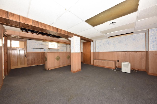 below grade area with a paneled ceiling, wood walls, dark carpet, and wainscoting