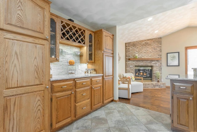 kitchen with light tile patterned floors, glass insert cabinets, light stone countertops, a brick fireplace, and vaulted ceiling