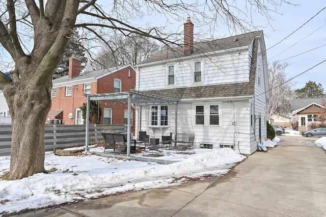 snow covered back of property featuring a shingled roof, fence, a gambrel roof, a chimney, and a pergola