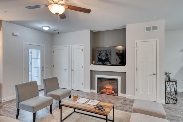 living room with visible vents, a ceiling fan, a warm lit fireplace, wood finished floors, and baseboards