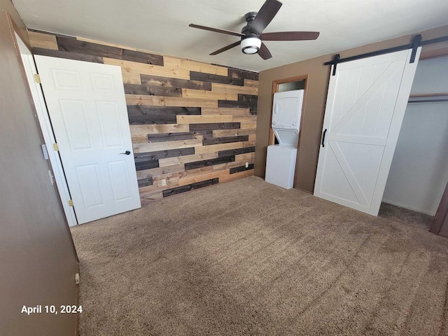 unfurnished bedroom with carpet, ceiling fan, wood walls, a barn door, and stacked washing maching and dryer