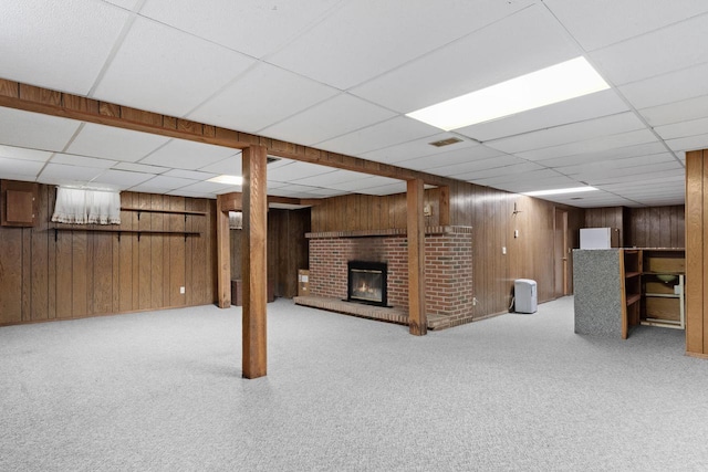 finished basement featuring a drop ceiling, a brick fireplace, wooden walls, and carpet floors