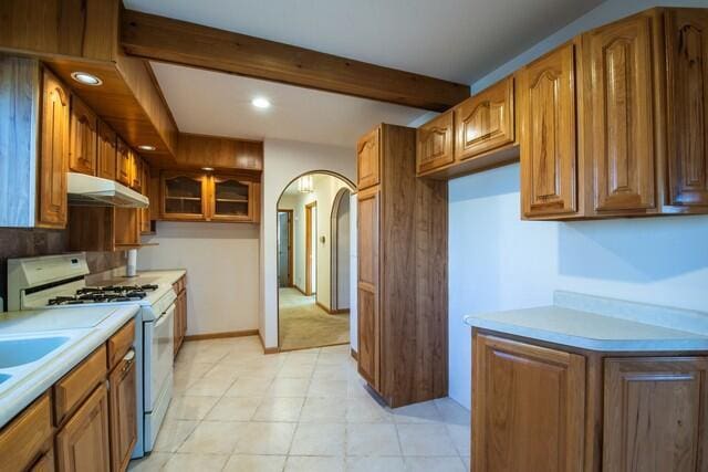 kitchen featuring beamed ceiling, under cabinet range hood, arched walkways, light countertops, and white gas range