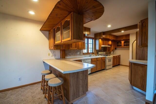 kitchen with glass insert cabinets, under cabinet range hood, a breakfast bar area, a peninsula, and white appliances