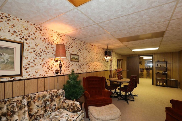 living area featuring wallpapered walls, a drop ceiling, a wainscoted wall, and carpet floors