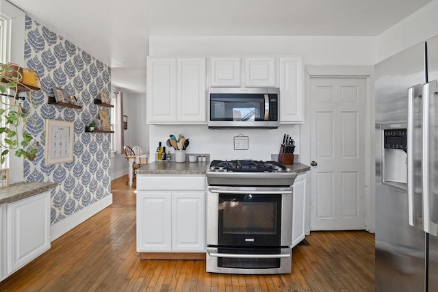 kitchen with light wood finished floors, wallpapered walls, baseboards, appliances with stainless steel finishes, and white cabinets