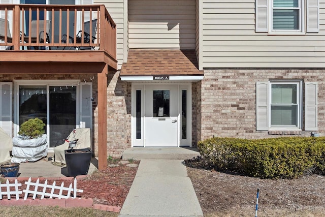 doorway to property featuring brick siding, roof with shingles, and a balcony