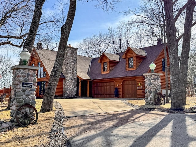 cabin featuring driveway, a chimney, an attached garage, and a shingled roof