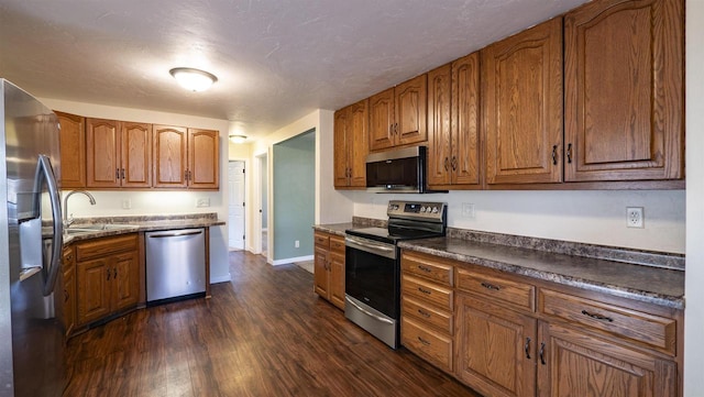 kitchen with dark countertops, dark wood finished floors, appliances with stainless steel finishes, brown cabinetry, and a sink