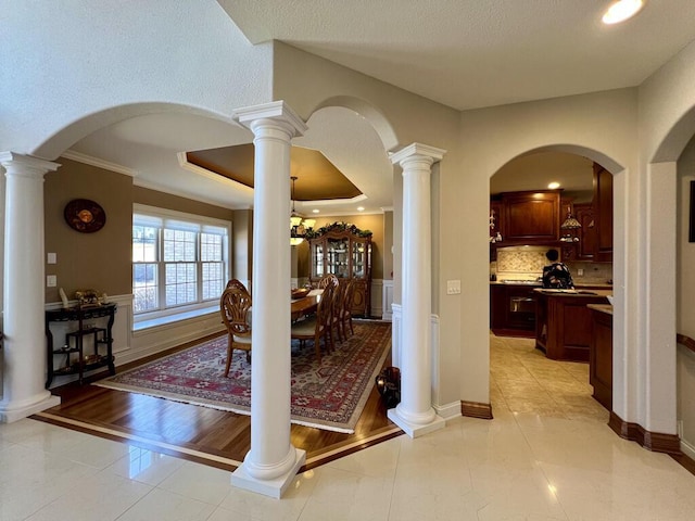 entryway featuring a raised ceiling, crown molding, light tile patterned floors, baseboards, and ornate columns