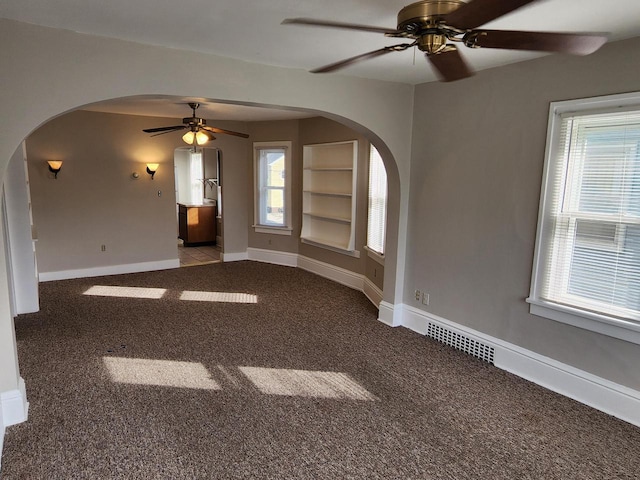 carpeted empty room featuring visible vents, built in shelves, arched walkways, baseboards, and ceiling fan