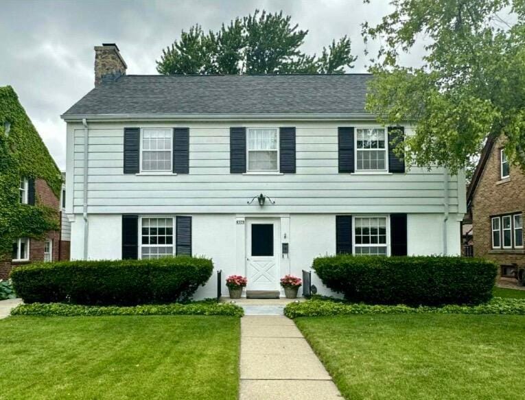 colonial home featuring a shingled roof, a front yard, stucco siding, and a chimney