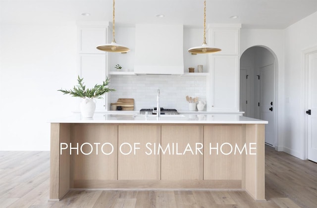 kitchen featuring tasteful backsplash, open shelves, custom range hood, white cabinetry, and a kitchen island with sink