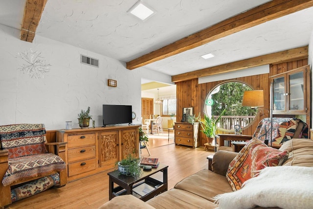 living room with wooden walls, visible vents, beamed ceiling, light wood-style floors, and a textured wall