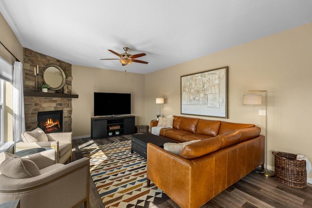 living area with baseboards, a ceiling fan, a stone fireplace, and dark wood-style flooring