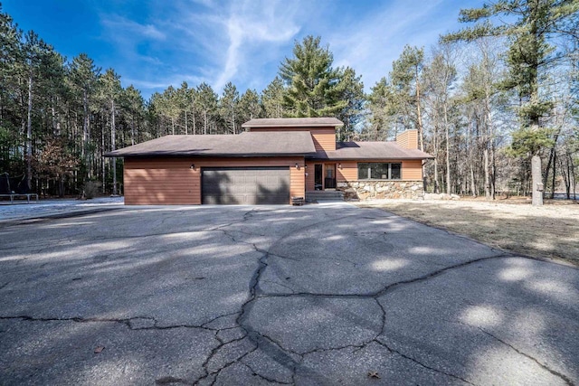 view of front of home with a wooded view, driveway, an attached garage, a chimney, and stone siding