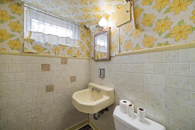 half bath featuring a wainscoted wall, toilet, a sink, tile walls, and wallpapered walls