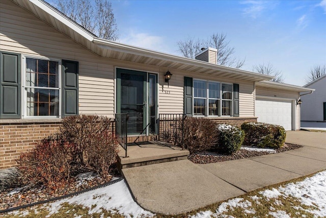 snow covered property entrance featuring concrete driveway, a garage, brick siding, and a chimney