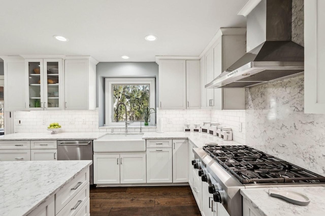 kitchen with a sink, light stone counters, appliances with stainless steel finishes, wall chimney range hood, and dark wood-style flooring