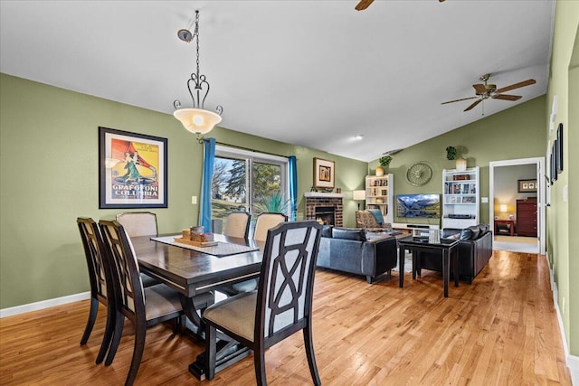 dining space featuring lofted ceiling, a brick fireplace, a ceiling fan, and light wood finished floors