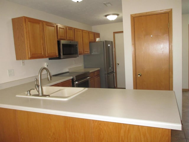 kitchen featuring brown cabinetry, a peninsula, a sink, stainless steel appliances, and light countertops