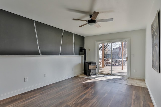 unfurnished room with a ceiling fan, a wood stove, baseboards, and wood finished floors