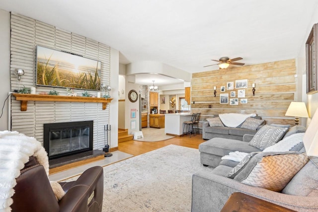 living area featuring light wood-style floors, a brick fireplace, wood walls, and ceiling fan with notable chandelier