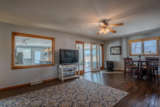 living room featuring visible vents, ceiling fan, baseboards, a wood stove, and dark wood-style flooring