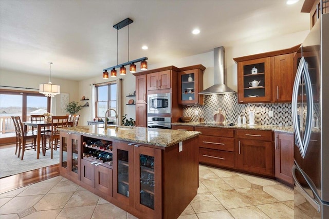 kitchen with wall chimney range hood, decorative backsplash, brown cabinets, appliances with stainless steel finishes, and a sink