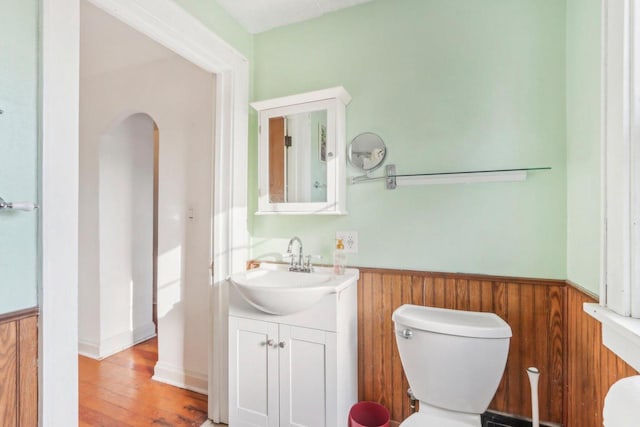 half bath with wooden walls, a wainscoted wall, toilet, wood finished floors, and vanity