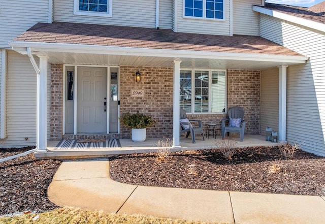 property entrance with brick siding, covered porch, and roof with shingles