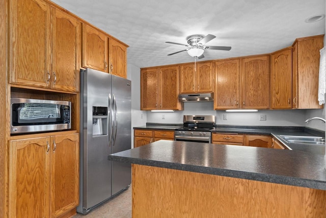 kitchen with a peninsula, a sink, under cabinet range hood, appliances with stainless steel finishes, and brown cabinets