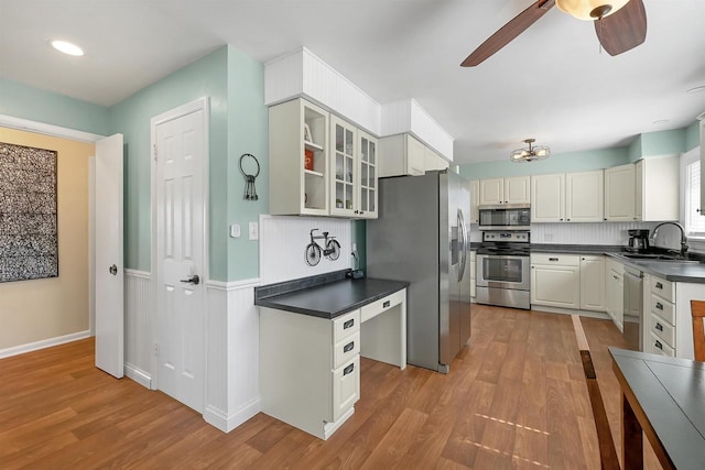 kitchen with a sink, dark countertops, light wood-style floors, appliances with stainless steel finishes, and glass insert cabinets